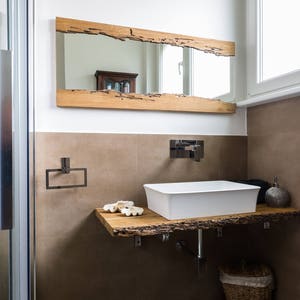 May include: A white porcelain bathroom sink with a wooden countertop and a rustic wooden mirror above it. The sink is in a modern bathroom with a shower and a towel rack.