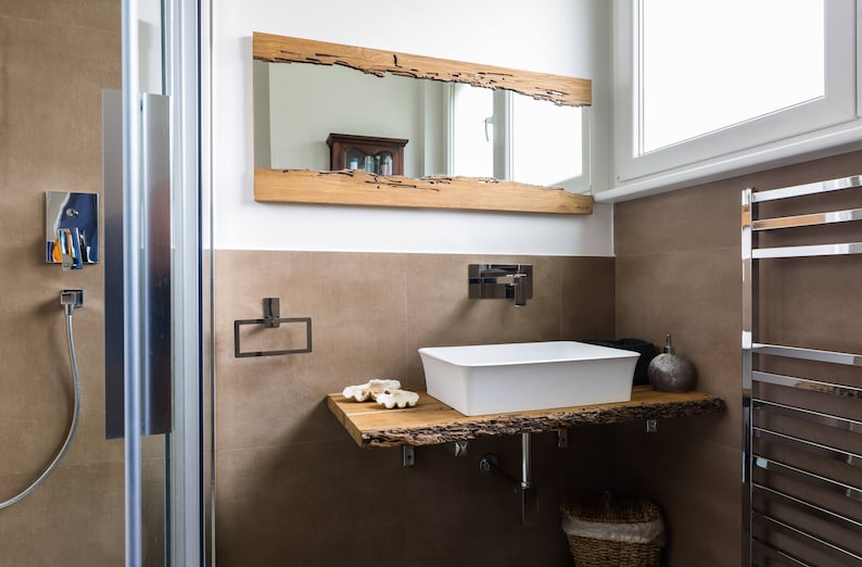 May include: A white porcelain bathroom sink with a wooden countertop and a rustic wooden mirror above it. The sink is in a modern bathroom with a shower and a towel rack.
