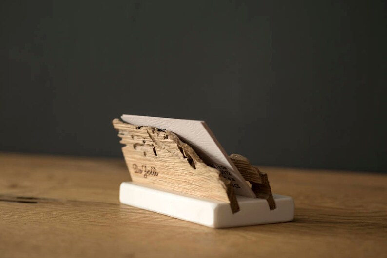 May include: A wooden business card holder with a natural wood finish. The holder is shaped like a mountain range and has a white base. The text "Del Solli" is engraved on the front of the holder.