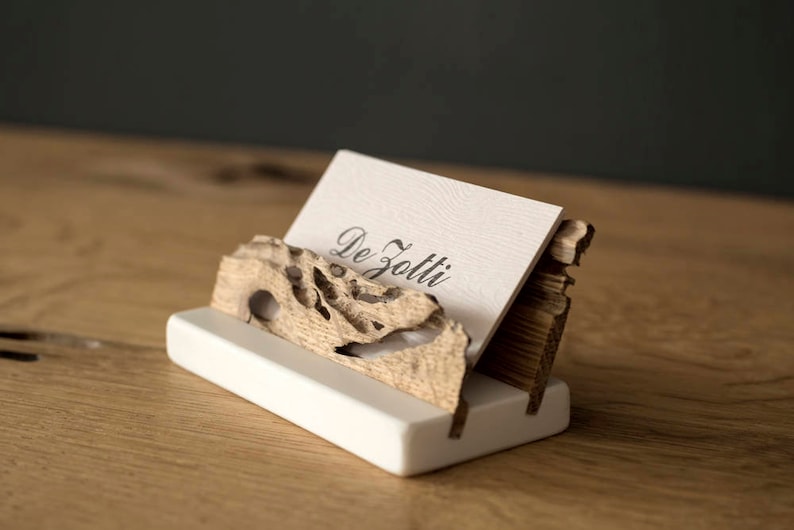 May include: A white ceramic business card holder with a wooden base. The base is made of a piece of driftwood with a natural, organic shape. The card holder is designed to hold a business card with the text "De Zotti" printed on it.