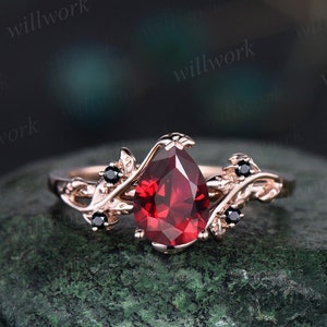 May include: A pear-shaped red gemstone ring set in rose gold with black diamond accents. The ring features a delicate leaf design.