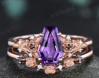 May include: A rose gold ring set featuring a central coffin-cut purple amethyst gemstone. The band is adorned with sculpted rose motifs, small clear accent stones, and tiny black round stones arranged in a floral-inspired design.