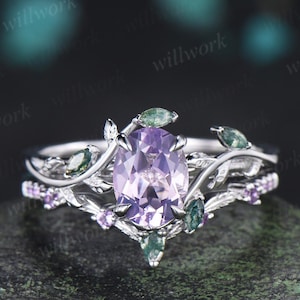 Peut inclure: Un ensemble de bagues en argent avec une pierre précieuse ovale violet clair au centre. La bague est ornée de détails délicats de feuilles et de vignes, et agrémentée de petites pierres précieuses vertes et violettes. La bague est posée sur une surface sombre et texturée.