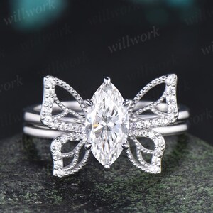 May include: A silver ring set featuring a marquise-cut diamond center stone. The ring's design includes a butterfly-shaped setting with pavé-set accent stones. The ring is displayed on a dark, textured surface.