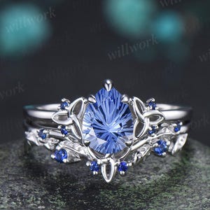 May include: A silver ring set featuring a pear-shaped, faceted blue gemstone as the centerpiece. The ring is surrounded by intricate floral and leaf designs, accented with smaller blue gemstones. The set includes a matching band.