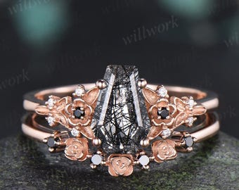 May include: A rose gold ring set featuring a central coffin-shaped rutilated quartz stone with black needle-like inclusions. The band is adorned with floral motifs, small clear accent stones, and tiny black round stones set in metal.