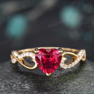 May include: A gold ring featuring a central heart-shaped red gemstone held by four prongs. The band is crafted with an intricate twisted design on both sides, accented with small, clear, round-cut stones embedded along the metal surface.