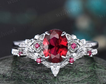 Puede incluir: Un anillo de compromiso de plata con un gran rubí ovalado rodeado de rubíes y diamantes más pequeños. El anillo presenta un diseño floral con delicadas hojas y flores.