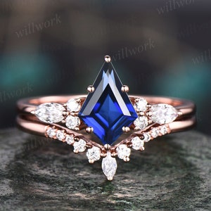May include: A rose gold engagement ring set with a blue sapphire center stone and a matching band with smaller diamonds. The sapphire is a square shape with pointed corners.
