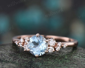 May include: A rose gold engagement ring with a light blue aquamarine center stone and a delicate halo of smaller diamonds. The band is adorned with a cluster of smaller diamonds on one side.
