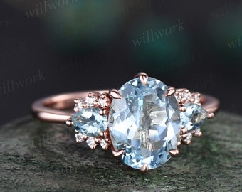 May include: A rose gold engagement ring with a large oval aquamarine center stone and two pear-shaped aquamarine side stones. The ring is set with small diamonds around the center stone and side stones.