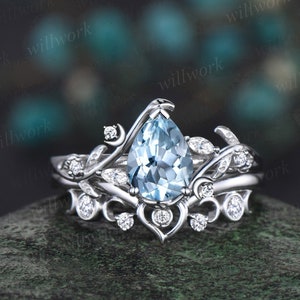 May include: A pear-shaped aquamarine engagement ring set in a delicate, floral-inspired design with a matching wedding band. The ring features a silver band with a pear-shaped aquamarine gemstone surrounded by smaller diamonds.
