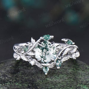 May include: A silver ring set featuring a central oval-cut gemstone with green inclusions, surrounded by smaller green stones and leaf-like details. The band has a delicate, nature-inspired design.