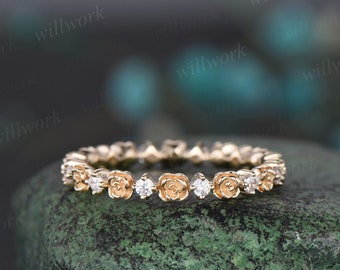 May include: A gold-colored ring featuring a floral design with small, round, clear gemstones. The ring has a repeating pattern of rose-shaped elements and sparkling stones. The ring is set against a textured, green surface.