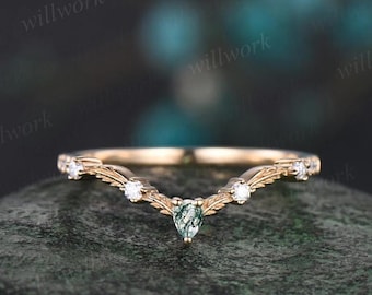 Op de afbeelding: Een delicate gouden ring met een gebogen ontwerp met een groene edelsteen en kleine heldere stenen. De ring ligt op een getextureerd oppervlak.