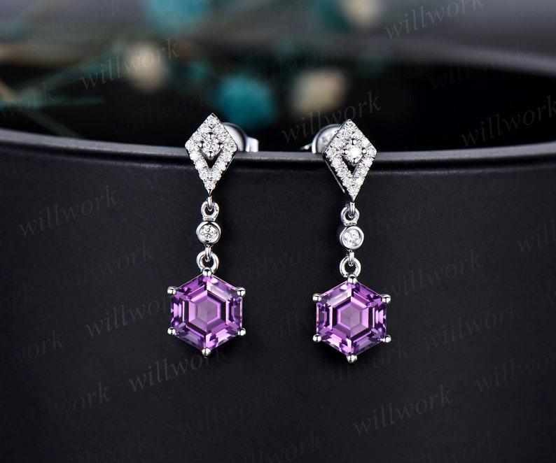 May include: A pair of dangle earrings with a hexagonal shaped purple gemstone and a diamond shaped setting with smaller diamonds surrounding the gemstone. The earrings are made of white metal.