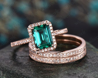 May include: A rose gold engagement ring set with a large emerald cut green gemstone surrounded by a halo of smaller diamonds. The set includes a matching wedding band with a row of diamonds.