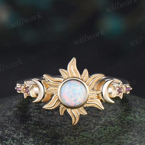 May include: A gold-toned ring featuring a central sunburst design with a white opal stone. Flanking the sun are crescent moons and small, purple gemstone accents. The ring has a delicate, celestial theme.