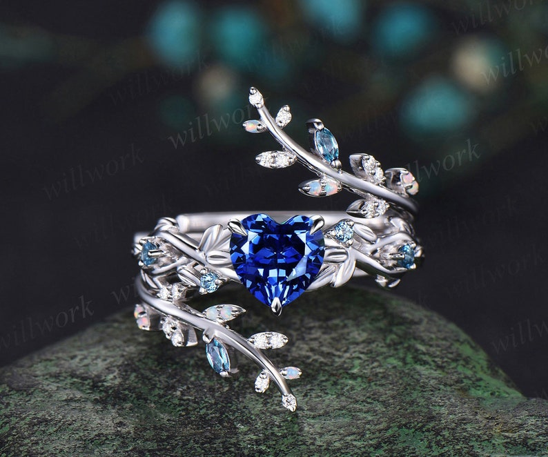 May include: A silver ring with a heart-shaped blue sapphire gemstone surrounded by smaller white and blue gemstones. The ring features a delicate, twisting vine design with leaves and small gemstones.