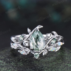 May include: A silver ring set featuring a pear-shaped moss agate center stone with a unique green pattern. The ring is adorned with small opals and leaf-like details, creating an elegant, nature-inspired design.