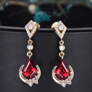 May include: A pair of gold earrings with a teardrop-shaped red gemstone and diamond accents. The earrings have a geometric design with a square-shaped top.