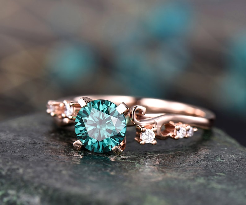 May include: A rose gold engagement ring with a large, round, teal green gemstone surrounded by smaller, round, white gemstones. The ring is set on a delicate band with a decorative, curved design.