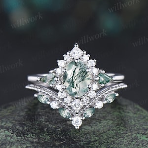 Peut inclure: Un ensemble de bagues de fiançailles en argent avec une pierre centrale en agate mousse verte de taille ovale, entourée de petits diamants. La bague comprend des accents de diamants supplémentaires et deux petites pierres vertes. La bague est posée sur une surface vert foncé.