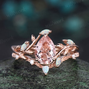 May include: A rose gold engagement ring with a pear-shaped morganite center stone and opal accents. The ring features a delicate floral design with leaves and branches.