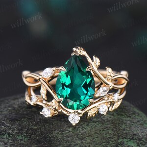 Puede incluir: Un anillo de compromiso de esmeralda en forma de pera engastado en una banda de oro con un delicado diseño de vid y pequeños diamantes. El anillo está sobre un fondo oscuro.