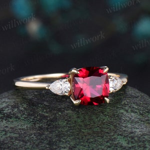 Puede incluir: Un anillo de oro con una gran piedra de rubí de forma cuadrada y dos diamantes más pequeños en forma de pera a cada lado.