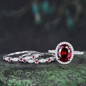 May include: A set of three rings. The engagement ring features a large oval-cut, deep red gemstone surrounded by a halo of smaller white stones. Two matching bands have alternating red and white stones. The rings are set on a green surface.