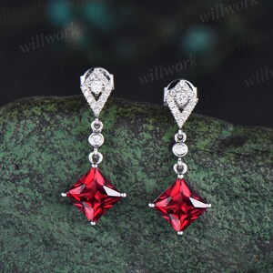 May include: A pair of dangle earrings with a square red gemstone and a diamond halo setting. The earrings are made of silver and have a drop design.