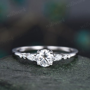May include: A silver engagement ring featuring a round centre stone and three pear-shaped accent stones. The ring is displayed against a grey backdrop.