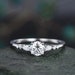 May include: A silver engagement ring featuring a round centre stone and three pear-shaped accent stones. The ring is displayed against a grey backdrop.