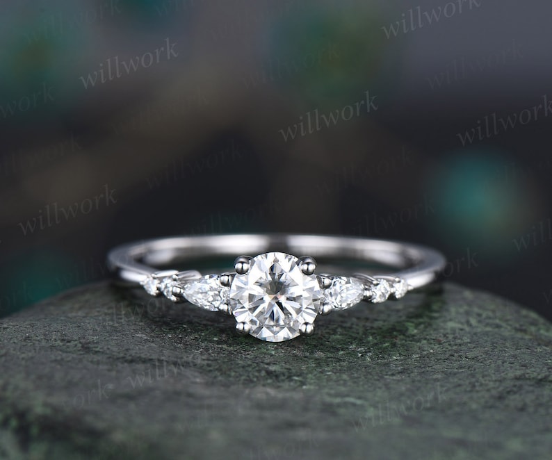 May include: A silver engagement ring featuring a round centre stone and three pear-shaped accent stones. The ring is displayed against a grey backdrop.