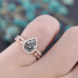 May include: Rose gold engagement ring set. The engagement ring features a pear-shaped, clear gemstone with black inclusions, surrounded by a halo of small, clear stones. The band is also adorned with small, clear stones. The wedding band has a matching design.