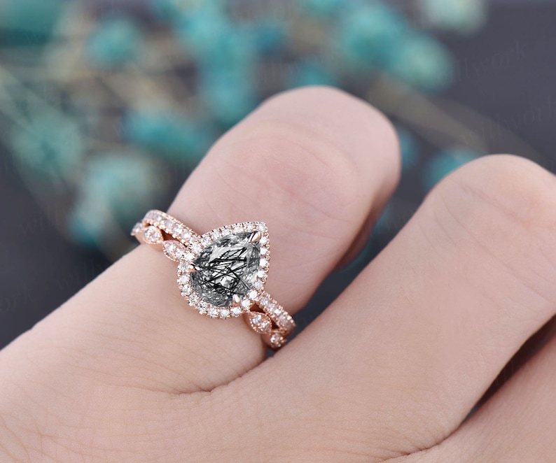 May include: Rose gold engagement ring set. The engagement ring features a pear-shaped, clear gemstone with black inclusions, surrounded by a halo of small, clear stones. The band is also adorned with small, clear stones. The wedding band has a matching design.