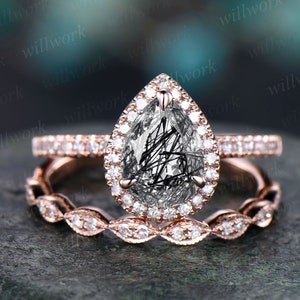 May include: A rose gold engagement ring set. The main ring has a pear-cut gemstone with black inclusions, encircled by a diamond halo. A matching band with a diamond design completes the set.