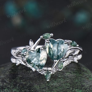 May include: A silver ring set featuring two heart-shaped gemstones with green and white patterns. The ring is adorned with small green accent stones and leaf-like details, creating an elegant and nature-inspired design.