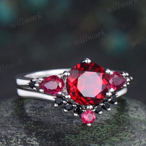 May include: A silver ring with a large, round, red gemstone as the centerpiece, flanked by two pear-shaped red stones and accented with smaller black and pink stones. The ring has a detailed, ornate design.