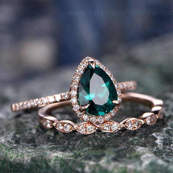May include: A pear-shaped emerald engagement ring set in rose gold with a halo of smaller diamonds. The ring is paired with a matching wedding band with a delicate design of smaller diamonds.