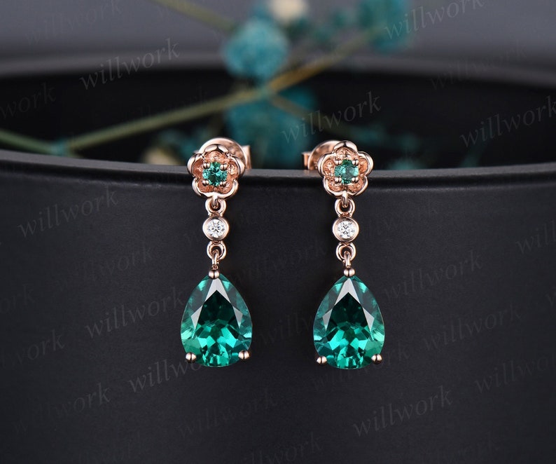 May include: A pair of dangle earrings with a pear-shaped emerald green gemstone and a small flower design. The earrings are made of rose gold and have a delicate design.