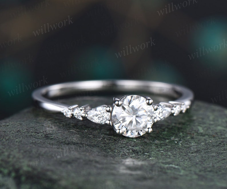 May include: A silver engagement ring featuring a round centre stone and pear-shaped accent stones. The ring is displayed against a grey background.