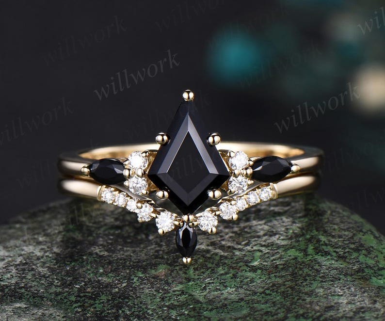 May include: A gold engagement ring with a black square-shaped center stone and smaller black stones on the band. The ring is surrounded by small white diamonds.