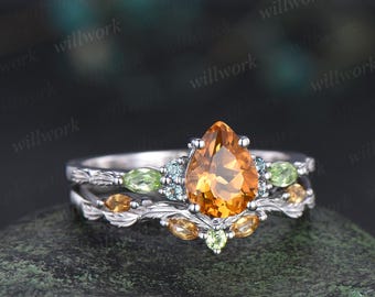 Könnte beinhalten: Silbernes Ring-Set mit einem birnenförmigen, bernsteinfarbenen Mittelstein. Der Ring ist mit kleinen, runden, grünen und blauen Edelsteinen sowie blattförmigen Details verziert.