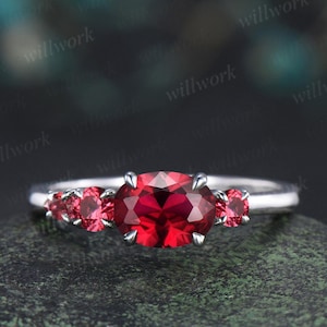 May include: A silver ring featuring a large, oval-cut, bright red gemstone as the centerpiece, flanked by two smaller red gemstones on each side. The ring is set against a dark, textured background.