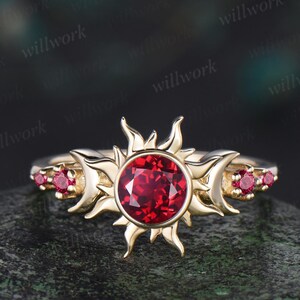 Könnte beinhalten: Ein goldener Ring mit einem roten Edelstein, der in einem Sonnenmotiv gefasst ist. Der Ring hat auf jeder Seite der Sonne eine Mondsichel und kleine rote Edelsteine auf der Ringschiene.
