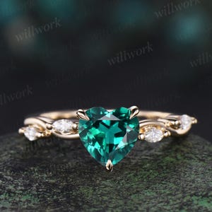 Peut inclure: Une bague en or avec une pierre précieuse verte émeraude en forme de cœur. La bague présente un anneau torsadé délicat avec de petites pierres d'accent claires. L'ensemble est posé sur un fond sombre et texturé.