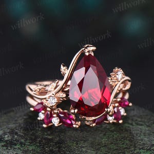 May include: A rose gold ring set featuring a large, pear-shaped, deep red gemstone as the centerpiece. The ring is adorned with smaller red gemstones and clear accent stones, with a floral design. The ring is set against a dark background.