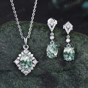 May include: A silver necklace and earrings set with a green moss agate stone pendant and matching earrings. The pendant and earrings are surrounded by small clear cubic zirconia stones.